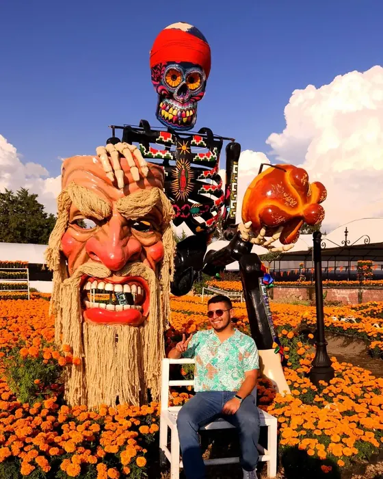 Rodolfo Gallegos sentado frente a una figura monumental de calavera y un campo de cempasúchil en Atlixco