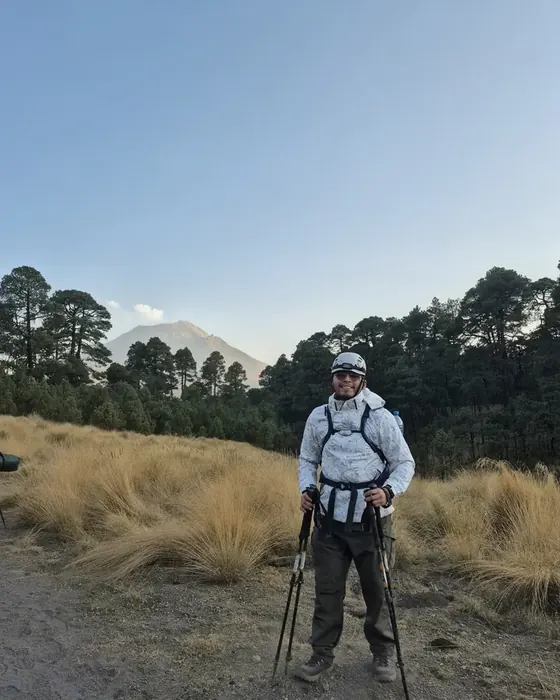Rodolfo Gallegos con equipo de montaña y bastones en un pastizal del Iztaccíhuatl, con el Popocatépetl detrás del bosque