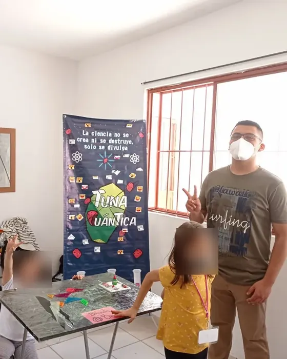 Rodolfo Gallegos con cubrebocas junto a una mesa de experimentos y una lona de divulgación científica, con dos niñas participando en el taller